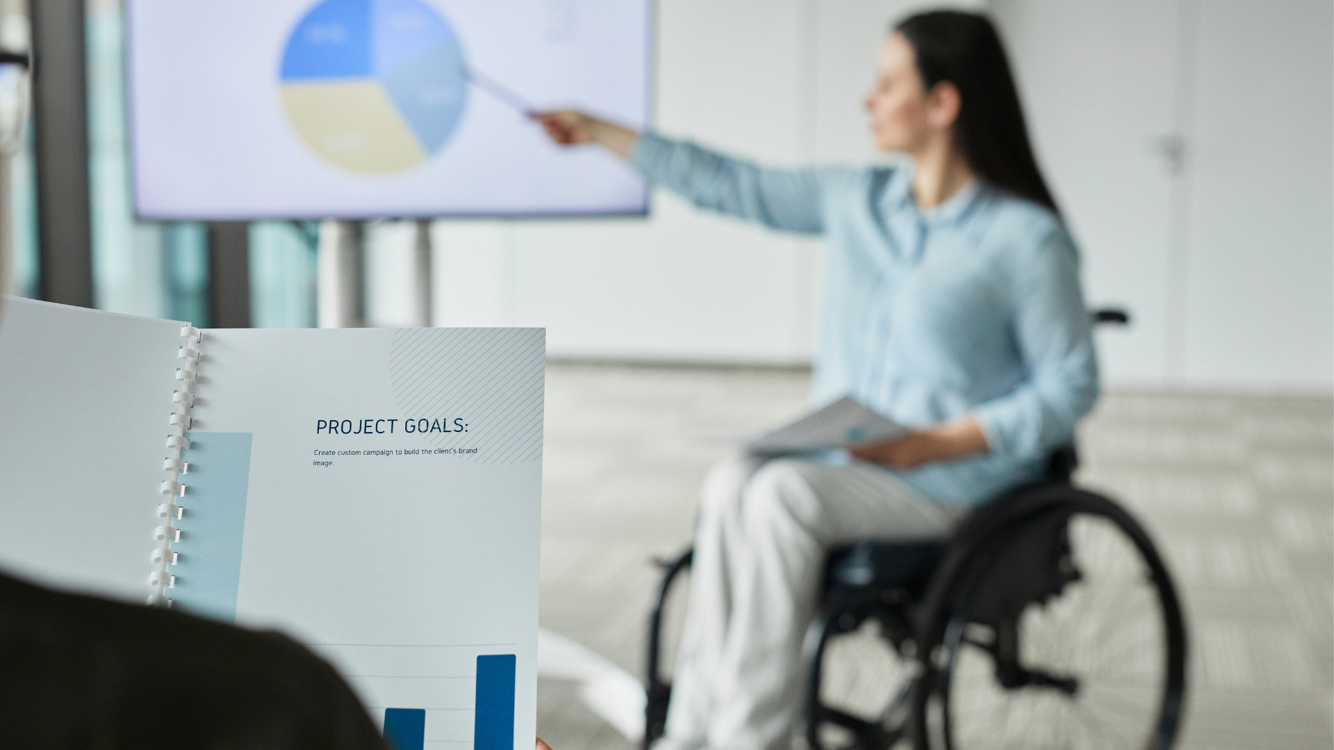 Osoba poruszająca się na wózku inwalidzkim prowadzi prezentację, wskazując na slajd z wykresem kołowym wyświetlony na ekranie. Na pierwszym planie widoczny jest fragment notesu z napisem „PROJECT GOALS”. W tle sala konferencyjna.