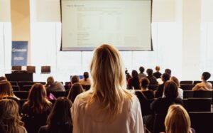 Zdjęcie przedstawia blondynkę stojącą na tle pełnej sali konferencyjnej.