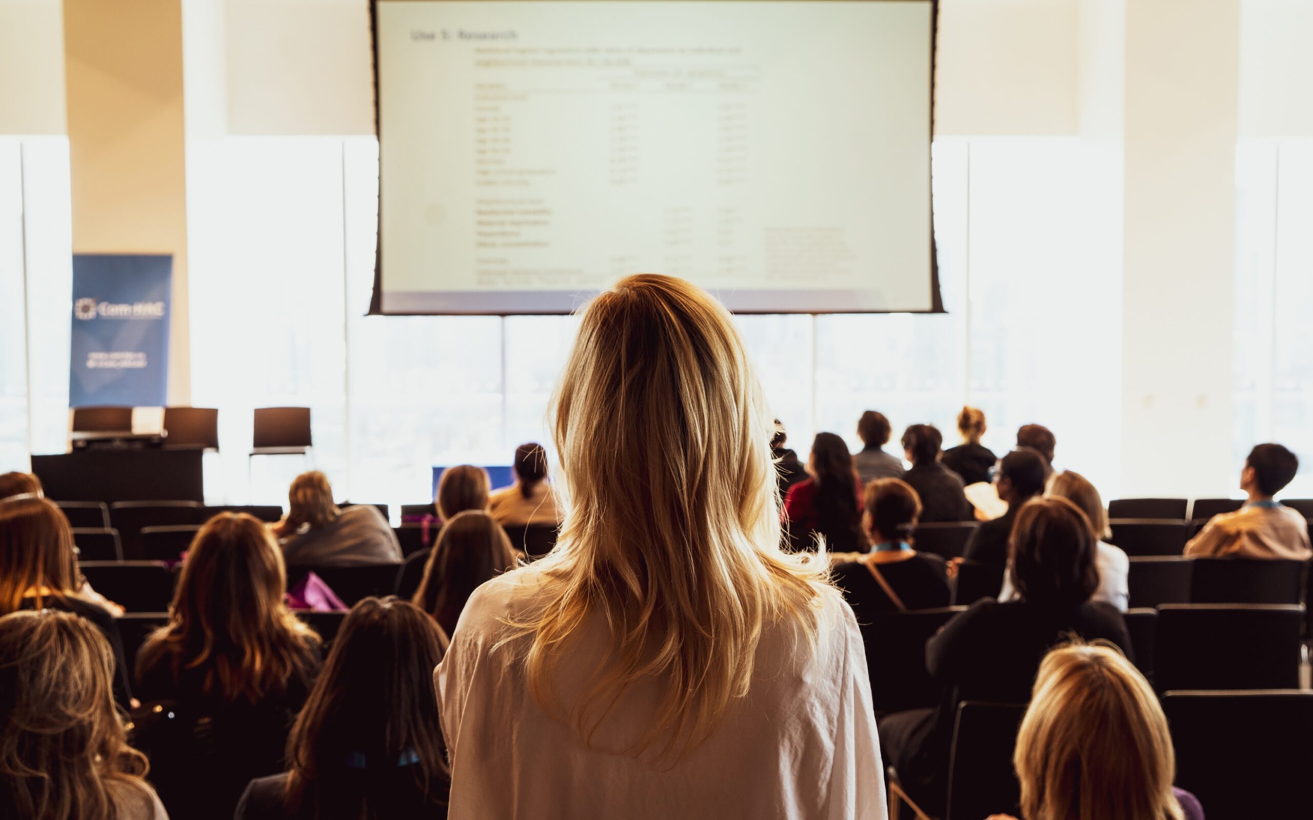 Zdjęcie przedstawia blondynkę stojącą na tle pełnej sali konferencyjnej.